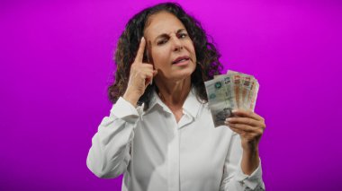 İngiliz banknotlarını ihtiyatlı bir şekilde mor arka planda tutan kadın, izole edilmiş renkli duvarlar üzerinde mali kararları düşünen orta yaşlı bir kadını gösteriyor..