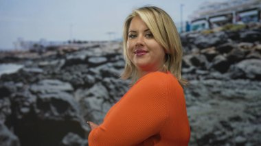 Turuncu kazaklı kadın güneşli sahilde kayalık sahile elini uzatırken açık gökyüzü altında gülümsüyor; memnuniyet.