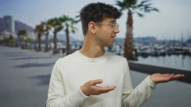 Beyaz süveter giyen ve gözlüklü adam, palmiyelerin palmiye ağaçlarının altında gezindiğini gösteriyor; belirsizlik..