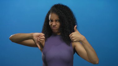 Mavi arka plana karşı karışık duyguları baş parmaklarıyla ifade eden kadın, kararsızlık veya şaşkınlık içinde, tecrit ortamında genç kadın çeşitliliğini vurguluyor.