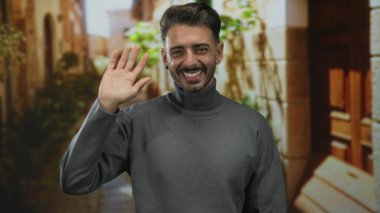 Sokakta el sallayan genç İspanyol adam, kaldırılmış avuç içi ve gülen yüz, taş duvarlar çerçeveli; hoş geldiniz.