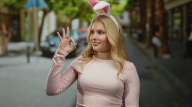 Sarı saçlı ve tavşan kulaklı gülümseyen kadın şehir sokaklarında şakacı hareketler yapıyor, açık havada neşeli şehir atmosferi sergiliyor..