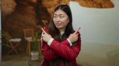 Kapalı bir yerde kırmızı ceketli çapraz kollarıyla poz veren bir kadın, rahat bir otel arka planına karşı kurulmuş, yön duygusunu ve karar verme mekanizmasını aktarıyor..