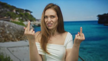 Kadın iki orta parmağını da parlak kumlu sahilde kayalık sahile doğru uzatıyor..
