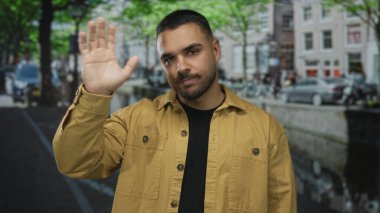 Sarı ceketli genç adam elini sallayarak caddede gözleri kapalı selam veriyor; sıcak karşılama.