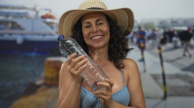 Güneşli limanda mayo giymiş, elinde şapkayla gülümseyen bir kadın. Arka planda tekneler, açık mavi gökyüzünü gösteren canlı bir deniz kenarı yaz gününü yakalıyor..