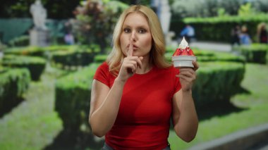 Parkta çilekli dondurmayı seven bir kadın, yeşilliklerle çevrili, kırmızı tişörtlü, sarı saçlı, arka planda heykeller olan, sakin bir yaz anı yaratan.