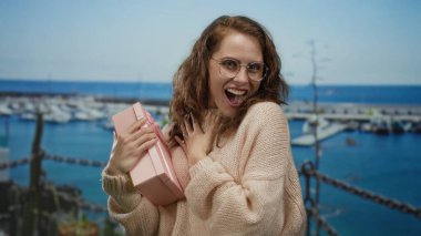 Sahilde pembe hediye kutusuyla gülümseyen, gözlüklü, süveterli, dışarıda duran, arka planda bulanık bir limanı olan bir kadın, mutluluğu işaret ediyor..
