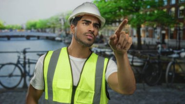 Sarı güvenlik yeleği ve beyaz şapkalı İspanyol adam inşaat halindeki binayı işaret ediyor..