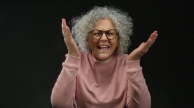 Gri saçlı, gözlüklü ve pembe gömlekli bir kadın. Duygusal olarak izole edilmiş, siyah bir arkaplan, anlamlı elleri ve ciddi bir yüz ifadesi var..