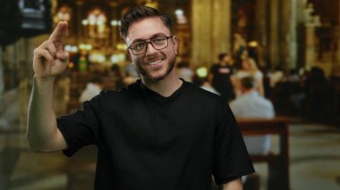 Kilisenin içinde sakallı genç adam jest yapıyor, gözlüklü ve siyah gömlekli, loş ışıklandırma altında sıralarda oturan insanların yumuşak arka planlarında güvenle gülümsüyor..