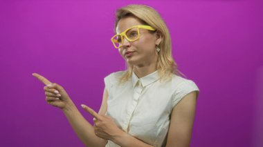 Sarı gözlüklü ve beyaz kolsuz bluzlu bir kadın mor bir stüdyoyu işaret ediyor..