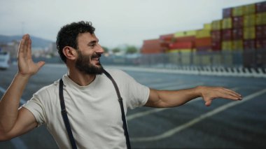 İspanyol sakallı bir adam arka planda gemi ve konteynırlarla bir limanda şakacı hareketler yapıyor. Deniz ve endüstriyel titreşimlerle canlı bir sahne yaratıyor..