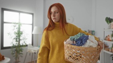 Çamaşır sepetini evdeki rahat çamaşır odasında tutan kadın. Pencereden süzülen güneş ışığı ve sakin atmosferi yükselten dekoratif bitkiler..