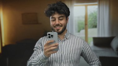 Samimi bir otel odasında akıllı telefon kullanan sakallı genç bir adam, arka plandaki pencere manzarasının yumuşak doğal ışıklandırmasıyla rahat bir atmosfer yaratıyor..