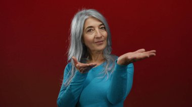 Gri saçlı, mavi kazaklı yaşlı kadın elini kırmızı duvara uzatıyor ve izole bir stüdyo çekiminde dostane bir gülümseme sergiliyor..