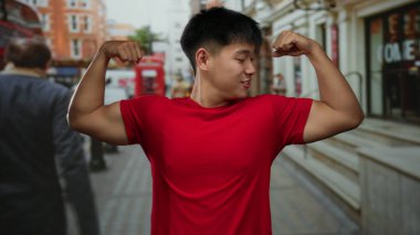 Kırmızı tişörtlü genç Çinli adam hareketli ve ikonik mimari ile dolu canlı bir şehir caddesinde kaslarını kasıyor..