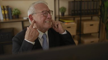 Takım elbiseli kıdemli bir adam ofiste oturup kulaklarını parmaklarıyla kapatarak iş yerindeki hayal kırıklığını ifade ediyor..