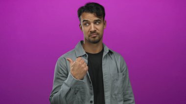 Gri ceketli genç ispanyol adam parlak pembe arka planda başparmak uçlarını işaret ediyor, kararsızlık ya da sorguyu kendine güvenen bir ifadeyle gösteriyor..