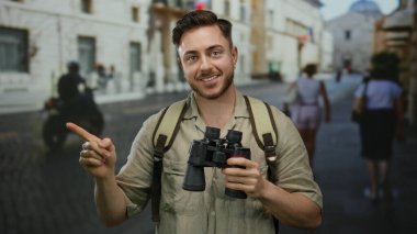 Sakallı ve dürbünlü genç adam güneşli bir günde şehir sokaklarında gezerken sırt çantasıyla gezerken gülümsüyor.