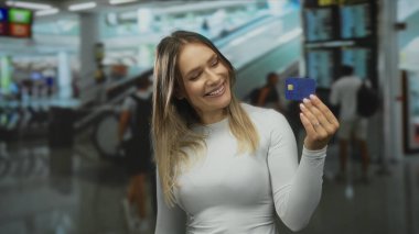 Havaalanında elinde mavi kredi kartıyla gülümseyen bir kadın. Sarışın, parlak ışıklarla çevrili ve arka planda bulanık gezginlerle..