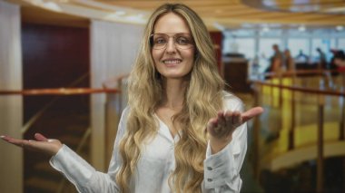 Modern bir otel odasında elleri açık el kol hareketi yapan, gözlüklü ve beyaz bluzlu sarışın bir kadın, kapalı mekandaki bulanık figürlerle çevrili..