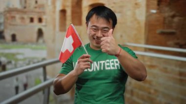 Kentsel alanda İsviçre bayrağı tutan, yeşil tişörtlü, gönüllü mesajlı, gülümseyen ve el hareketi yapan, gündüz vakti sokakta duran bir adam..