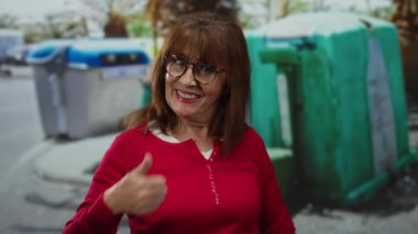 Kırmızı kazaklı, gülümseyen İspanyol kadın şehir sokaklarında renkli geri dönüşüm kutularının yanında, kentsel çevresel farkındalığın altını çiziyor..