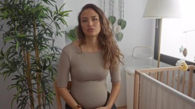 Uzun saçlı hamile bir kadın, yumuşak dekor ve doğal ışıkla çevrili, rahat bir bebek odasında oturur. Huzuru ve annelik beklentisini somutlaştırır..
