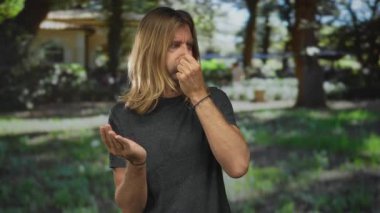 Uzun sarı saçlı bir adam burnunu sıkar ve uzun ağaçların altındaki yeşil parkta bir şeyi koklamak için avuç içini açar..