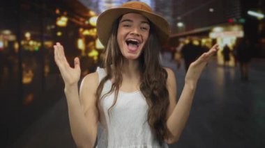 Hasır şapka ve beyaz elbise giyen genç İspanyol kadın, yoğun şehir caddesinde geceleri omuz silkiyor; iyimserlik.