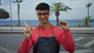 Gözlüklü ve önlüklü genç adam deniz kenarında palmiye ağaçları ve okyanus manzaralı bir yerde sessizlik jesti yapıyor.