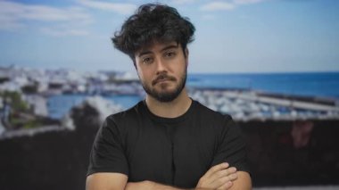 Sakallı genç adam limana yakın bir yerde duruyor. Arkaplanı açık gökyüzünün altında, gündüzleri siyah tişört giyerken.