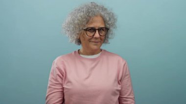 Gri saçlı, gözlüklü ve pembe kazaklı yaşlı kadın mavi arka planda şakacı hareketler yapıyor..