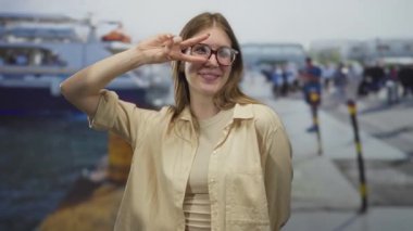 Gözlüklü sarışın kadın gülümsüyor ve limanda demirli teknelerin yanında barış işareti yapıyor. Mutluluk..