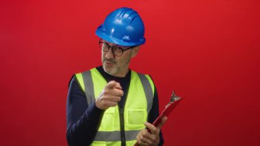 Miğferi ve yeleği olan adam, kırmızı arka plana karşı pano tutuyor. İnşaat sektöründe kararlılık ve güveni gösteriyor.