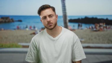 Sakallı genç adam arka planda deniz ve palmiye ağaçları olan bir sahilde duruyor, rahat bir deniz kenarı manzarası yakalıyor..