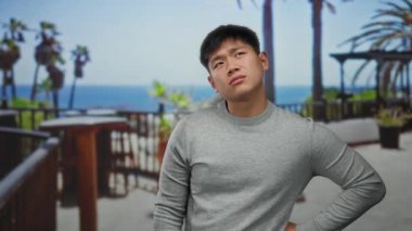 Gri kazaklı genç adam, güneşli bir günde derin düşüncelere dalmış, palmiye ağaçları ve okyanus manzaralı deniz kıyısında geziniyor..