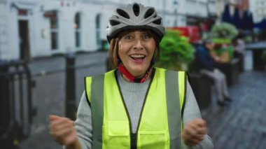 Kasklı ve yansıtıcı yelek giyen yaşlı İspanyol kadın canlı bir şehir caddesinde neşeli jestler yaparak canlı şehir atmosferini yakalıyor..