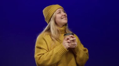 Elinde kahve fincanı tutan, sarı kazak ve şapkalı, mavi arka planda gülümseyen bir kadın..