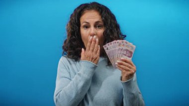 Orta yaşlı bir kadın, izole edilmiş mavi arka plana karşı sürpriz bir ifadeyle Meksika pezosu tutuyor..