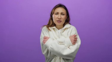 Kadın canlı bir mor arka plan duvarına karşı soğuk bir şekilde soyutlanmış hissediyor, rahat beyaz bir kazak giyiyor, kollarını sıkıca kavuşturup rahatsızlık veriyor..