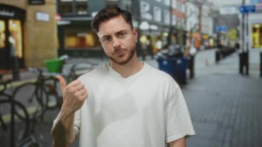 Beyaz tişörtlü, sakallı genç adam şehir caddesinde başparmak kaldırıyor. Etrafı dükkan ve bisikletlerle çevrili..