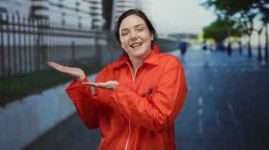 Turuncu tulumlu kadın dışarıda gülümsüyor ve arka planda çitle bir şehir caddesinde el kol hareketi yapıyor. Ya özgürlük ya da kaçış öneriyor..