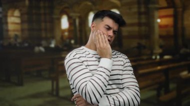 Kilisede çizgili tişört giyen genç bir adam. Düşünceli bir yüz ifadesi var. Kapalı alanda sakin bir ortam yaratıyor..