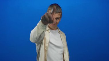 Kafkasyalı bir adam, izole edilmiş, mavi bir arkaplan önünde sırıtarak kamerayı işaret ediyor..