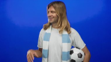 Hayran eşarbı takan sarışın adam futbol topunu kolunun altında tutuyor ve stüdyoda el hareketi gösteriyor; coşku.