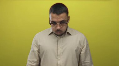 Stüdyoda elini kaldırmış bej gömlekli ve gözlüklü İspanyol genç adam sarı arka planda özgüven ve odaklanma.