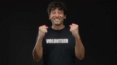 Gönüllü tişört giyen İspanyol bir adam, siyah bir arkaplan karşısında heyecan gösteriyor, çeşitli jestler yoluyla coşku gösteriyor..