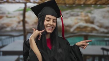 Mezuniyet şapkalı ve cüppeli genç İspanyol kadın terasta gülümserken telefon jesti yapıyor..